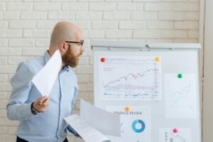 Bald businessman in smart casual attire analyzing financial charts on a whiteboard in an office setting.